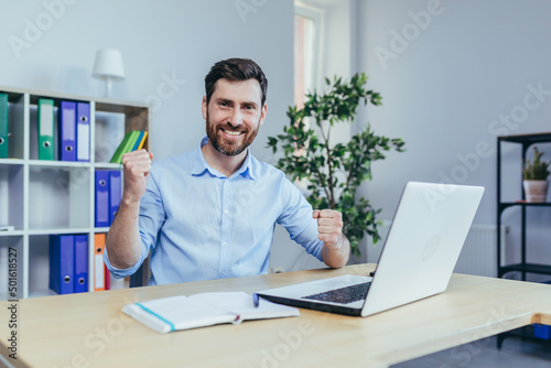 Successful businessman with a beard looks at the camera and smiles, celebrates triumph, works on a laptop in the office