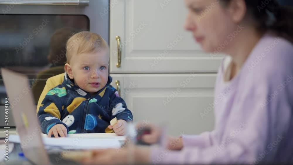 Working woman with one year old baby boy in high chair, mother can free her hands to go about her business. Mother with child works on laptop at home. High quality 4k footage Working woman with one year old baby boy in high chair, mother can free her hands to go about her business. Mother with child works on laptop at home. High quality 4k footage