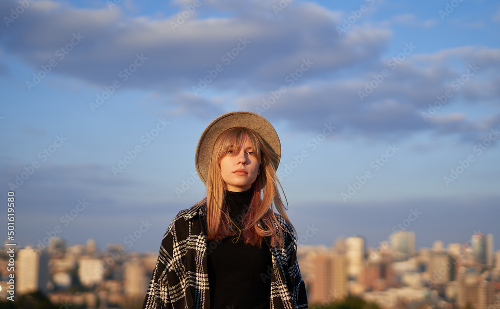 Attractive glamour blonde long hair woman in stylish clothes - shirt and brown hat stands on the background of blue cloudy sky and urban city background. Online shopping concept. High quality image