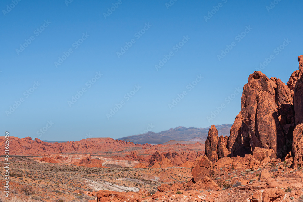 Overton, Nevada, USA - February 24, 2010: Valley of Fire. Wide red rock landscape under blue sky with mountain range on horizon and dry desert floor with shrubs around. 