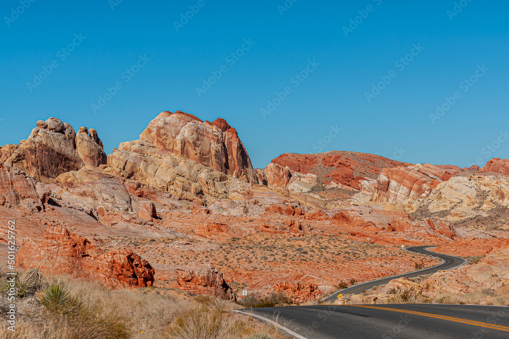 Fototapeta premium Overton, Nevada, USA - February 24, 2010: Valley of Fire. Up and down goes the black asphalt road with yellow divider between beige-red rock formations under blue sky over dry desert floor.