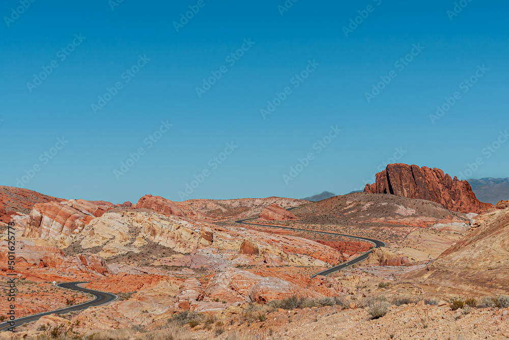 Naklejka premium Overton, Nevada, USA - February 24, 2010: Valley of Fire. Long meandering black asphalt road meandering through very wide landscape of red and beige rock over dry desert floor under blue sky.