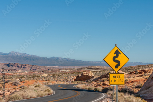 Overton, Nevada, USA - February 24, 2010: Valley of Fire. Black on yellow next-4-miles meandering sign along black asphalt road in wide desert landscape under light blue sky and mountains on horizon.