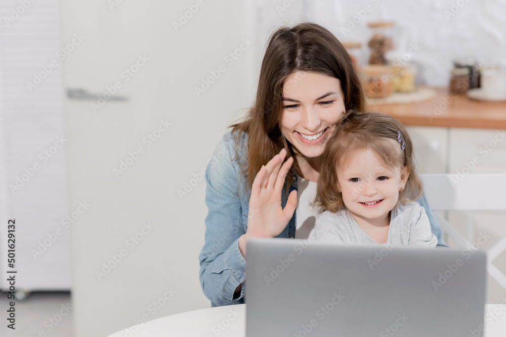 Happy young woman with joyful toddler having video call from relatives, waving hello to them. Beautiful mother with little daughter smiling, using computer to talk online with family while quarantine Happy young woman with joyful toddler having video call from relatives, waving hello to them. Beautiful mother with little daughter smiling, using computer to talk online with family while quarantine
