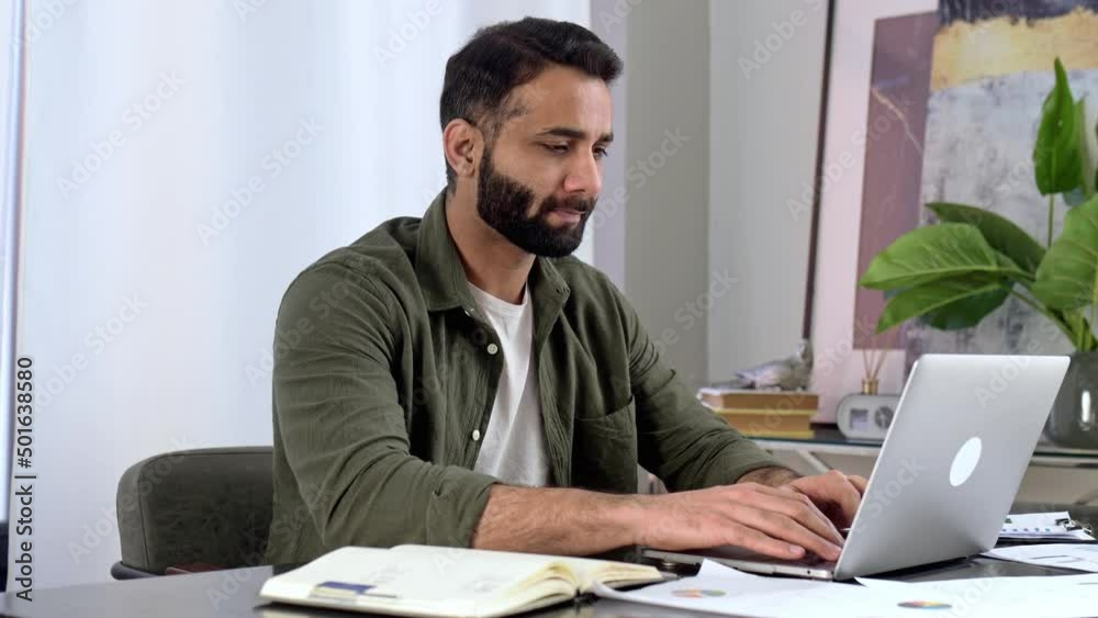 Positive smart indian or arabian man in casual clothes, freelancer or manager, sitting at work desk in the office, listening to webinar, taking notes, learning, improving skills, smiling Positive smart indian or arabian man in casual clothes, freelancer or manager, sitting at work desk in the office, listening to webinar, taking notes, learning, improving skills, smiling