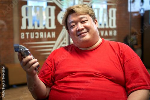 Smiling fat barber with a hair clipper on the hand while sitting on a barber shop © DavidMorillo