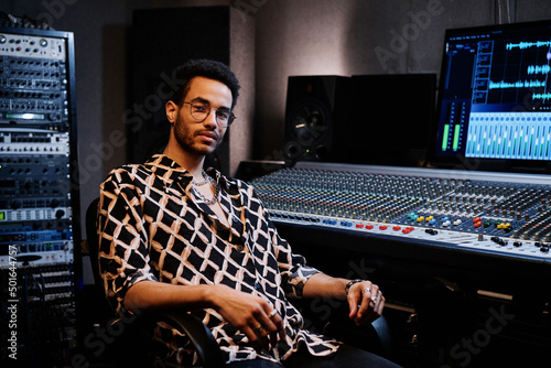 Young African American recording producer wearing fashionable outfit sitting against mixing console in studio photo
