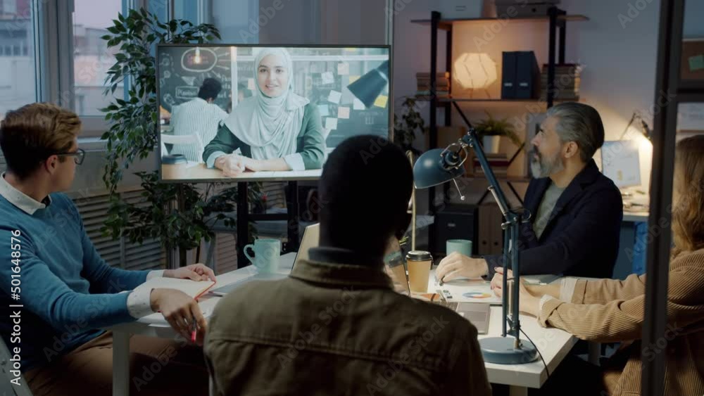 Diverse group of employees speaking to muslim business partner during online meeting in dark office. Communication and technology concept. Diverse group of employees speaking to muslim business partner during online meeting in dark office. Communication and technology concept.