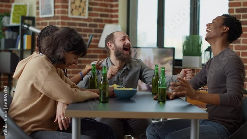Happy friends enjoying gathering with beer and snacks, having conversation and laughing together. Group of positive people drinking beer and making jokes in living room. Leisure activity.