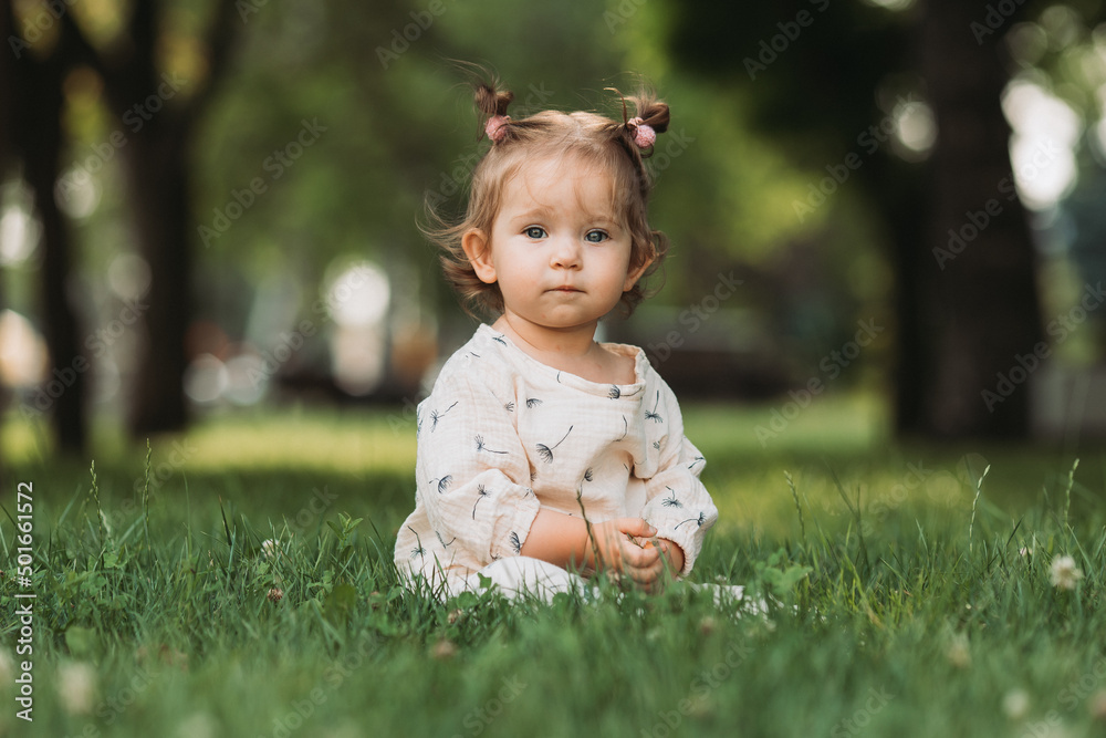 cute little girl with a funny hairstyle is sitting on a blooming green lawn in the park. lifestyle cute little girl with a funny hairstyle is sitting on a blooming green lawn in the park. lifestyle