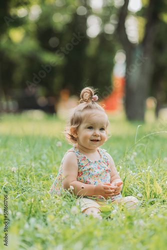 cute little girl with a funny hairstyle is sitting on a blooming green lawn in the park. lifestyle
