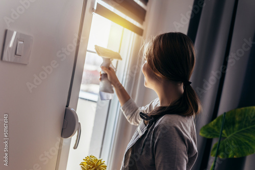 Happy Beautiful Girl Cleaning Window by spraying Cleaning Products and wiping with Sponge.
