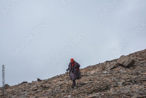 Tourist on stone hill at very high altitude under gray cloudy sky. Hiker with trekking poles and camera on rocky mountain in overcast weather. Man in high mountains among sharp rocks and sharp stones.