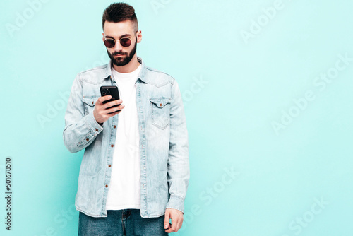 Handsome confident model.Sexy stylish man dressed in jacket and jeans. Fashion hipster male posing near blue wall in studio. Holding smartphone. Looking at cellphone screen. Using apps