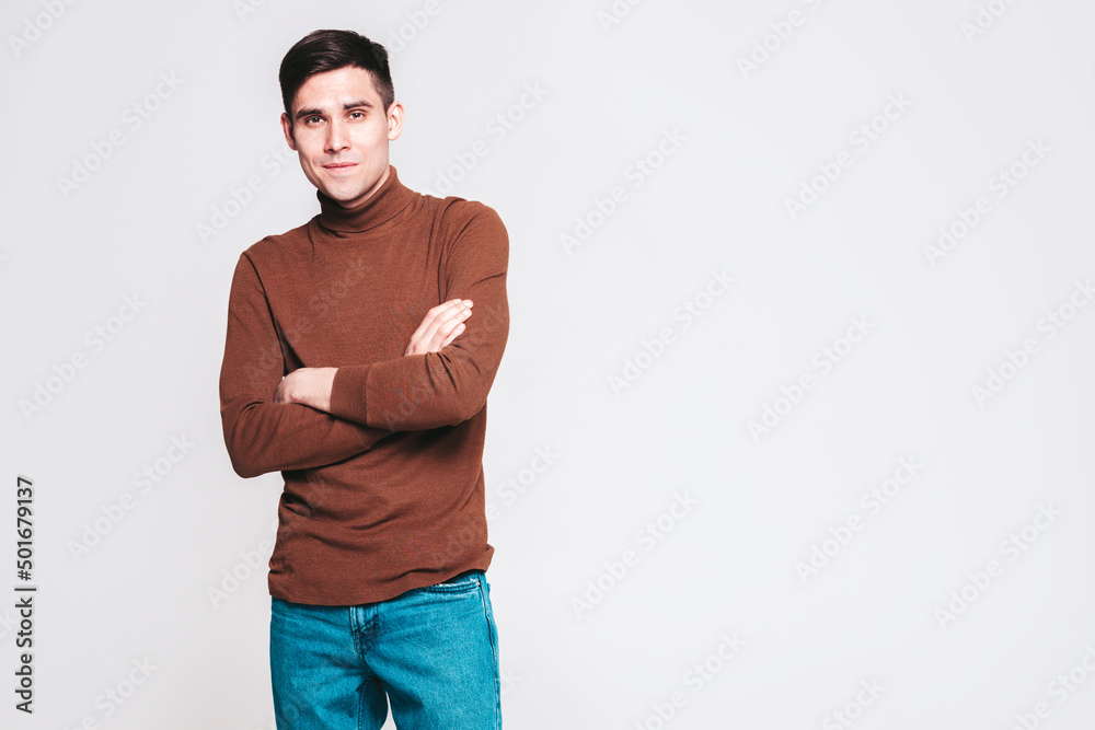 Portrait of handsome smiling model. Sexy stylish man dressed in brown turtleneck sweater and jeans. Fashion hipster male posing on grey background in studio Portrait of handsome smiling model. Sexy stylish man dressed in brown turtleneck sweater and jeans. Fashion hipster male posing on grey background in studio