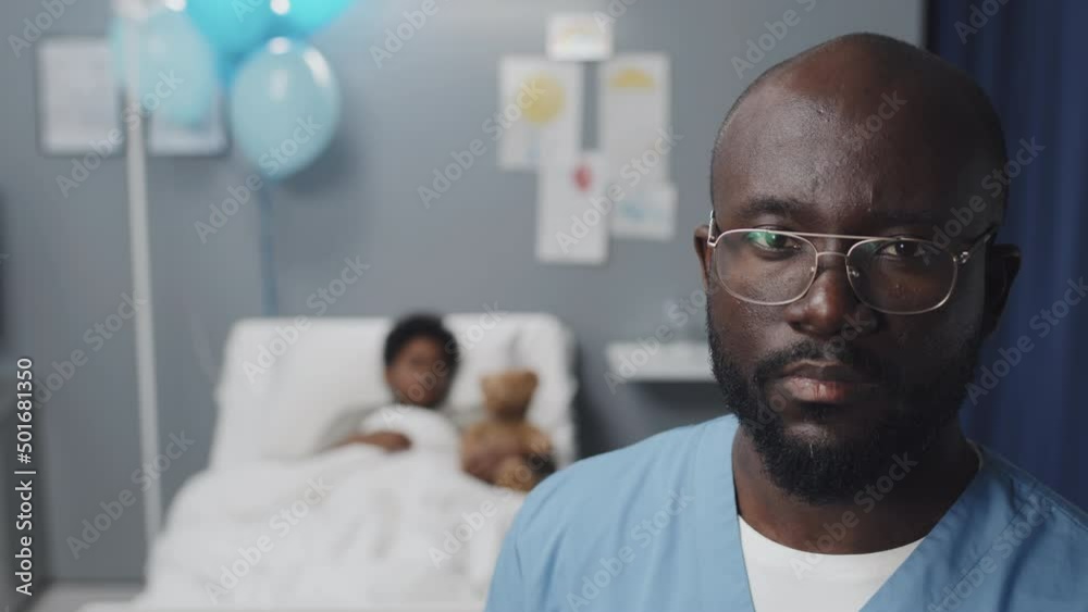 Shoulder-up of young African American male nurse wearing scrub suit and eyeglasses standing in hospital ward of blurred kid who lying in bed, man looking on camera