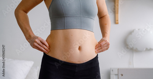Close up of a belly with scar from c-section and side belly fat. Women’s health. A woman dressed up in sportswear demonstrating her imperfect body after a childbirth with nursery on the background. photo
