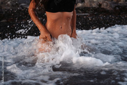 slender body of a sexy girl erotically sitting on her knees in the water in waves of sea on beach in summer on vacation. Small blur from a long exposure