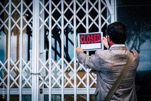 Businesswoman closing her business activity due economy crisis putting closed sign on the door of his store. Concept of bankrupt and shotdown commercial job occupation. Back view of man
