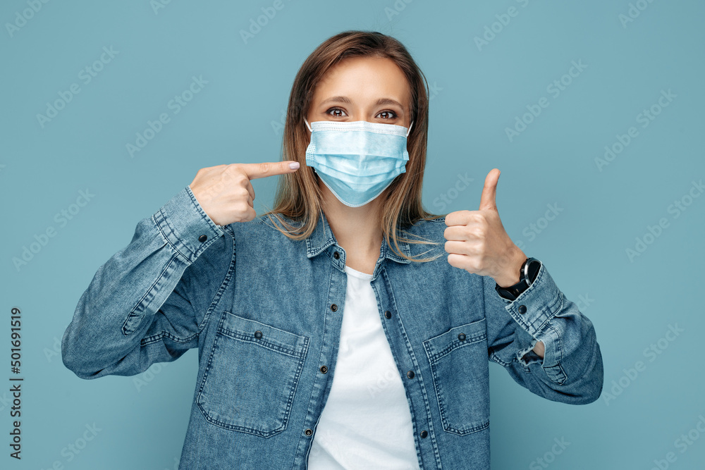 Young woman pointing finger on medical mask on blue background. Female showing thumb up. Young woman pointing finger on medical mask on blue background. Female showing thumb up.