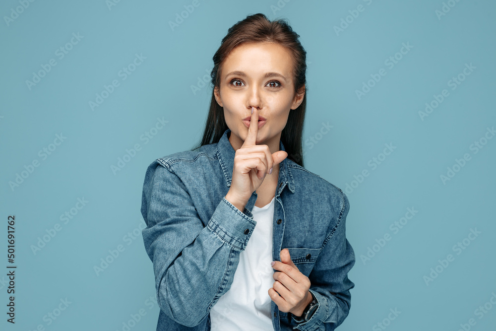 Young woman in jeans shirt showing keep silent gesture looking to camera over blue background. Young woman in jeans shirt showing keep silent gesture looking to camera over blue background.
