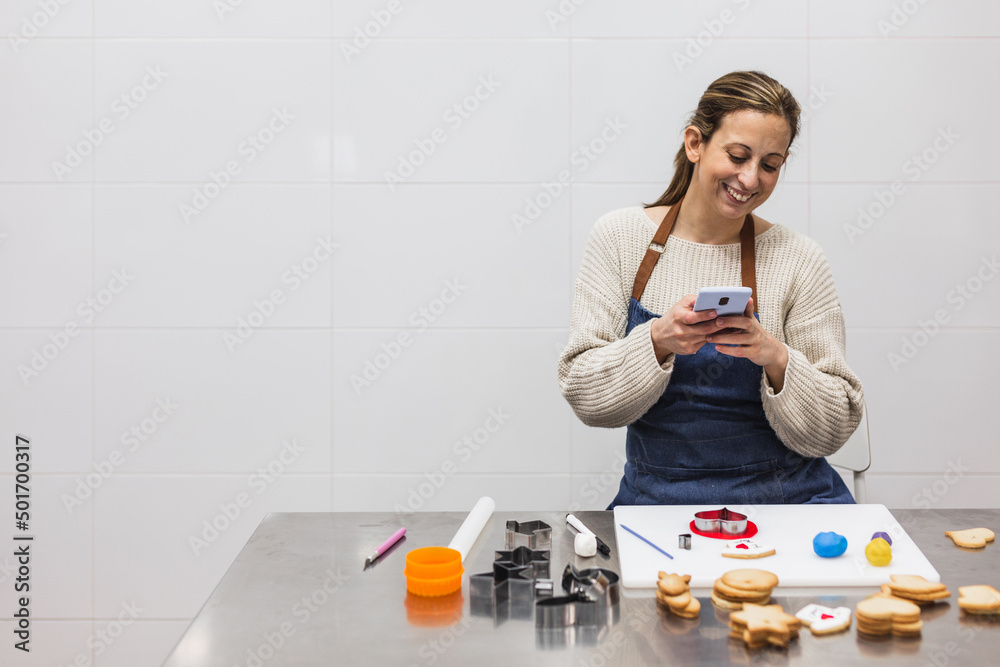 Smiling pastry chef using her mobile phone while working in the kitchen of the pastry shop. Smiling pastry chef using her mobile phone while working in the kitchen of the pastry shop.