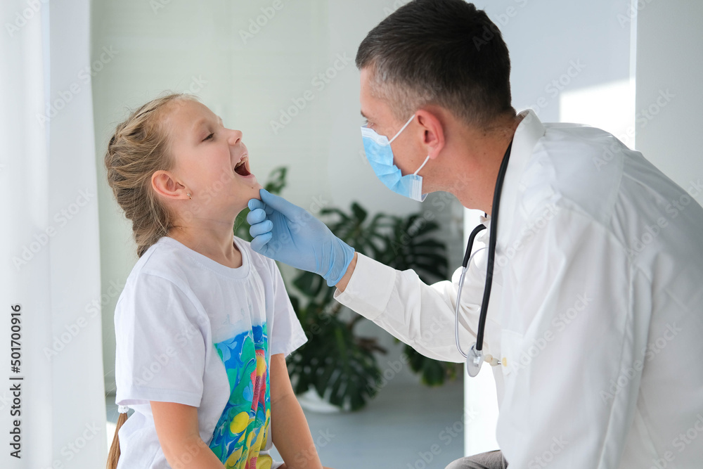 Obraz premium A doctor in a medical mask examines the throat of a child. A doctor examines a little girl during an illness. Calling a doctor at home.