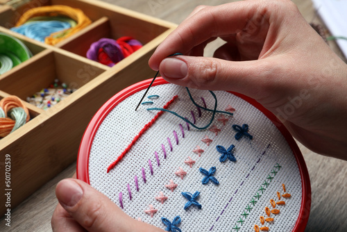 Woman doing different stitches with colorful threads on fabric in embroidery hoop at wooden table, closeup photo