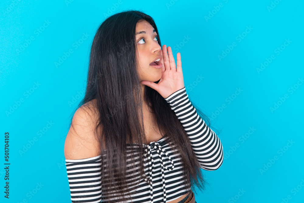 young latin woman wearing striped T-shirt over blue background profile view, looking happy and excited, shouting and calling to copy space. young latin woman wearing striped T-shirt over blue background profile view, looking happy and excited, shouting and calling to copy space.