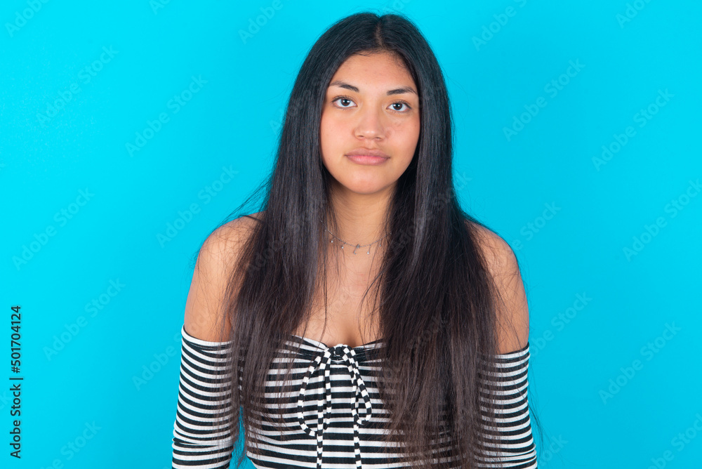Fototapeta premium Joyful young latin woman wearing striped T-shirt over blue background looking to the camera, thinking about something. Both arms down, neutral facial expression.