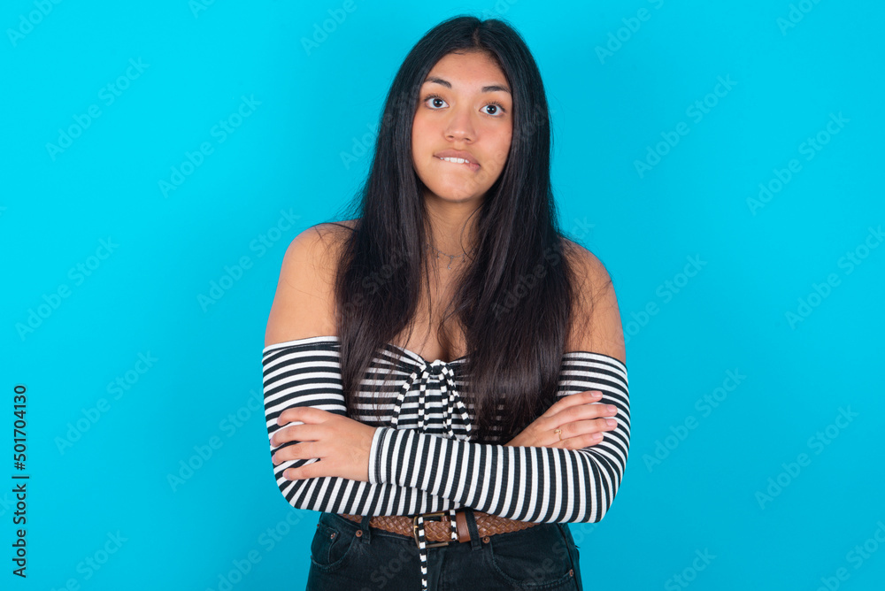 young latin woman wearing striped T-shirt over blue background bitting his mouth and looking worried and scared crossing arms, worry and doubt. young latin woman wearing striped T-shirt over blue background bitting his mouth and looking worried and scared crossing arms, worry and doubt.