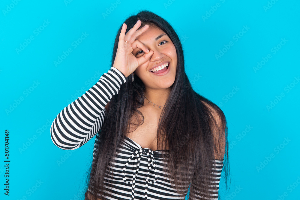 young latin woman wearing striped T-shirt over blue background with happy face smiling doing ok sign with hand on eye looking through finger. young latin woman wearing striped T-shirt over blue background with happy face smiling doing ok sign with hand on eye looking through finger.