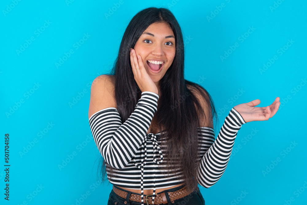 Positive glad young latin woman wearing striped T-shirt over blue background says: wow how exciting it is, indicates something.  One hand on his head and pointing with other hand.
