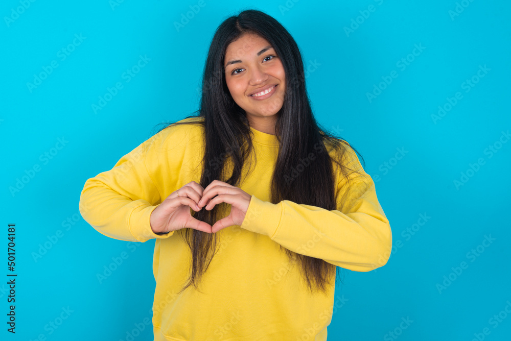 young latin woman wearing yellow sweater over blue background smiling in love showing heart symbol and shape with hands. Romantic concept. young latin woman wearing yellow sweater over blue background smiling in love showing heart symbol and shape with hands. Romantic concept.