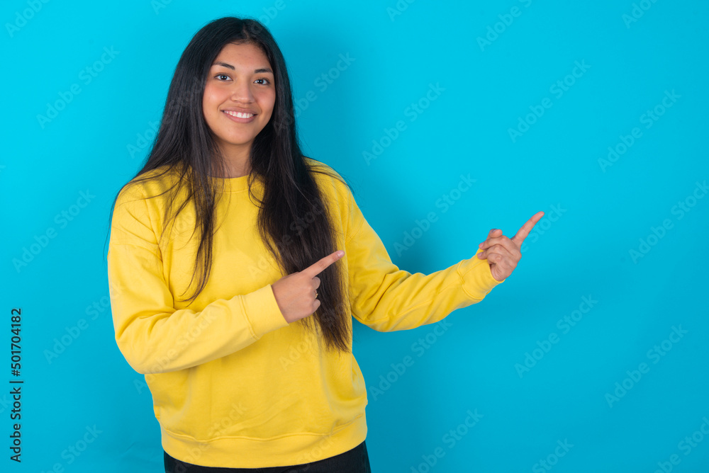 young latin woman wearing yellow sweater over blue background with positive expression, indicates with fore finger at blank copy space for your promotional text or advertisement. young latin woman wearing yellow sweater over blue background with positive expression, indicates with fore finger at blank copy space for your promotional text or advertisement.