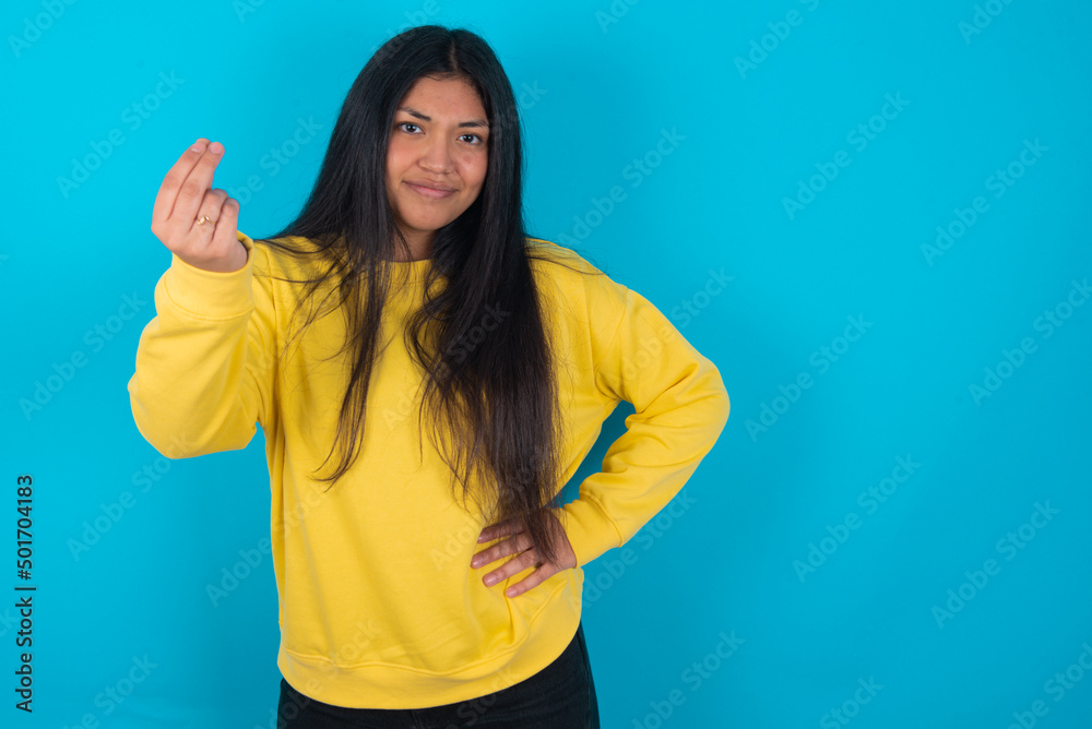 Fototapeta premium What the hell are you talking about. Shot of frustrated young latin woman wearing yellow sweater over blue gesturing with raised hand doing Italian gesture, frowning, being displeased and confused 