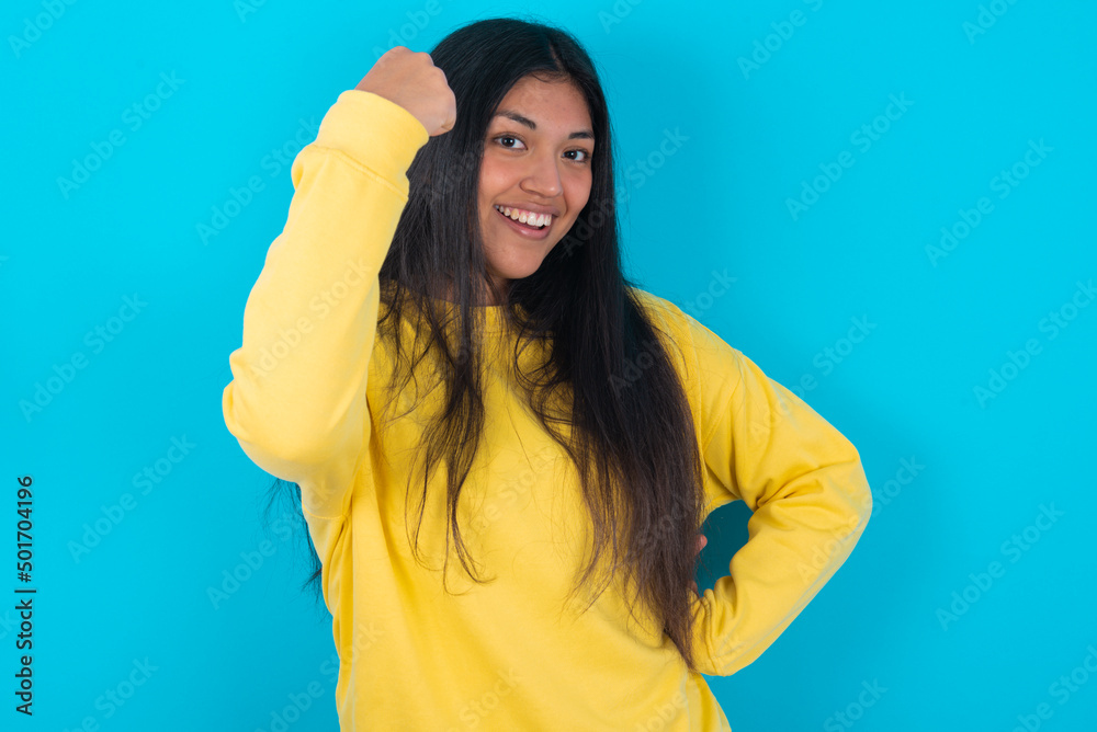 Overjoyed young latin woman wearing yellow sweater over blue background glad to receive good news, clenching fist and making winning gesture. Overjoyed young latin woman wearing yellow sweater over blue background glad to receive good news, clenching fist and making winning gesture.