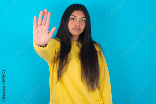 young latin woman wearing yellow sweater over blue background doing stop gesture with palm of the hand. Warning expression with negative and serious gesture on the face. © Jihan
