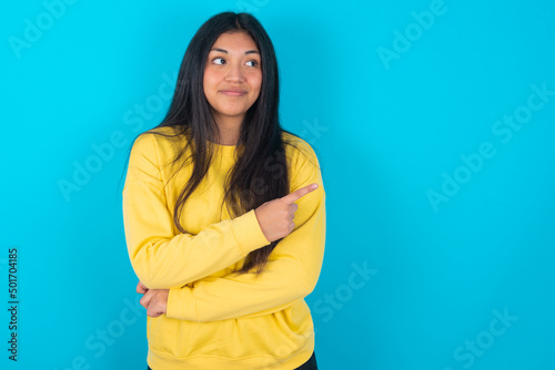 Portrait of young latin woman wearing yellow sweater over blue background posing on camera with tricky look, presenting product with index finger. Advertisement concept.