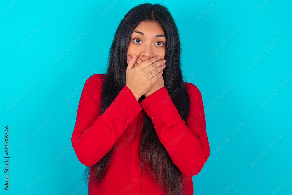 Stunned young latin woman wearing red shirt over blue background covers mouth with both hands being afraid from something or after hearing stunning gossips. Stunned young latin woman wearing red shirt over blue background covers mouth with both hands being afraid from something or after hearing stunning gossips.