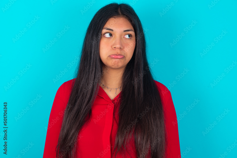 Dissatisfied young latin woman wearing red shirt over blue background purses lips and has unhappy expression looks away stands offended. Depressed frustrated model.