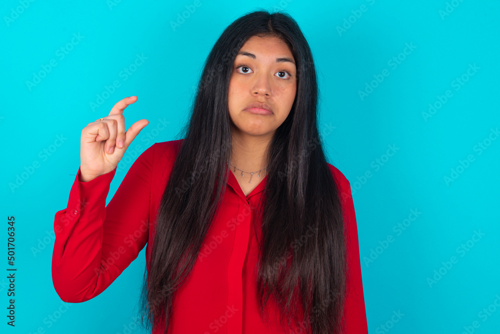 young latin woman wearing red shirt over blue background purses lip and gestures with hand, shows something very little. young latin woman wearing red shirt over blue background purses lip and gestures with hand, shows something very little.