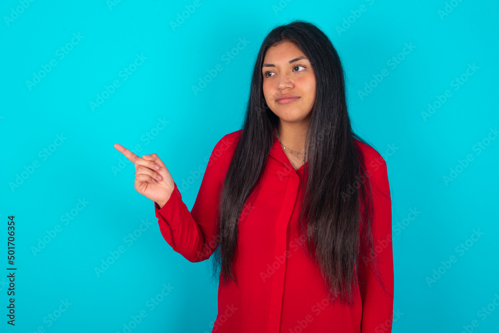 young latin woman wearing red shirt over blue background points at copy space and advertises something, advices best price.