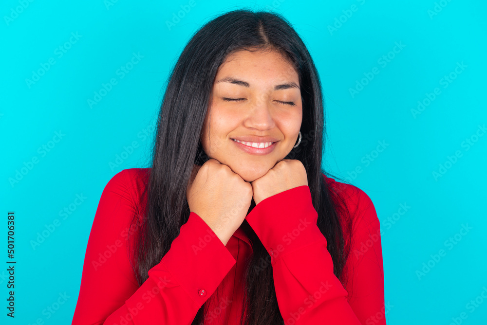 Cheerful young latin woman wearing red shirt over blue background has shy satisfied expression, smiles broadly, shows white teeth, People emotions Cheerful young latin woman wearing red shirt over blue background has shy satisfied expression, smiles broadly, shows white teeth, People emotions