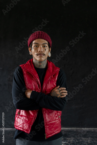 Portrait of cool Asian hip hop young bearded man with tattoo on face, neck and hands standing and cross his arms with nose piercing, big ear piercing and wear red knit hat and vest. Black background