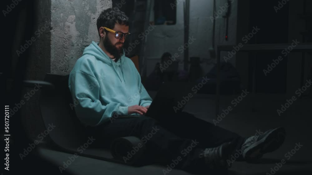 Handheld shot of male IT developer working with software and website at night on a laptop during the war in a dark bomb shelter