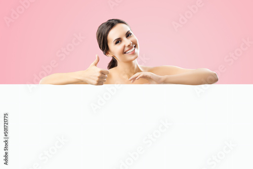 Excited smiling brunette woman with naked shoulders  undress  showing thump up hand sign gesture  standing behind white banner mock up signboard with copy space area  over rose pink color background.