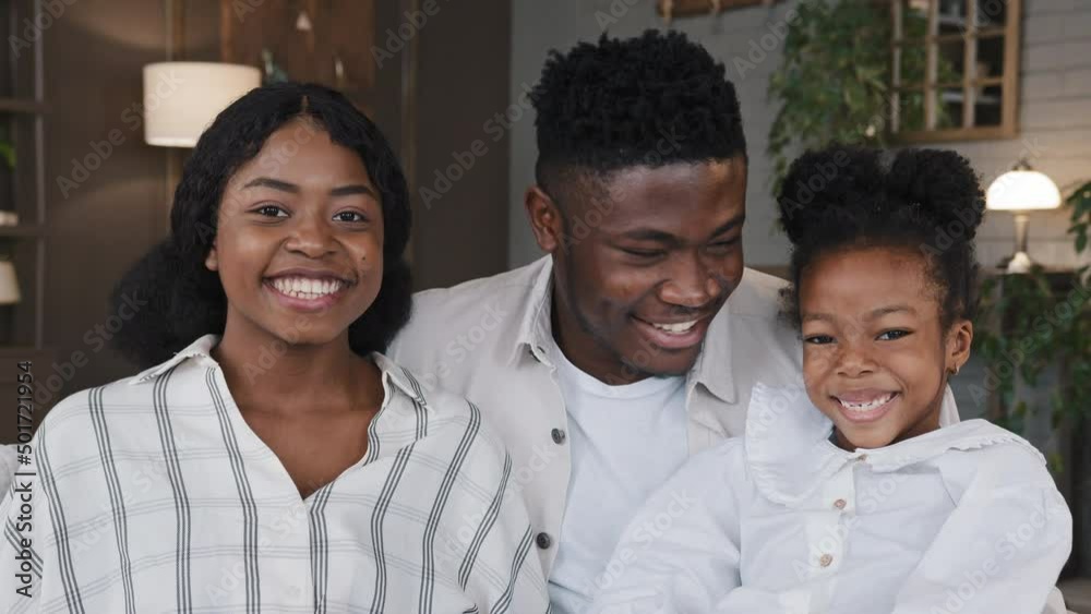 Happy African family portrait. Parents with cheerful daughter girl looking at camera laughing cuddling at home smiling toothy. Positive mother, father and child hugging on sofa. Dental health concept Happy African family portrait. Parents with cheerful daughter girl looking at camera laughing cuddling at home smiling toothy. Positive mother, father and child hugging on sofa. Dental health concept