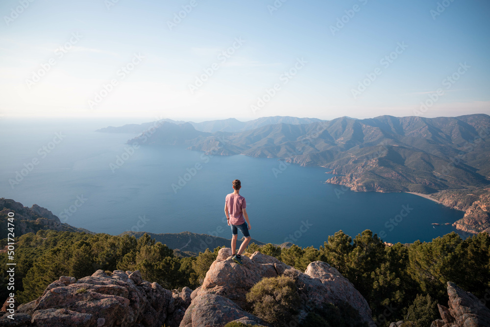 Man standing alone on an isolated rock in the Calanques de Piana and looking at the landscape, in Corsica - Panoramic view