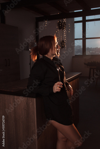 teen leisure. young girl in black long shirt stands casual in profile with long spoon in hand and looks apart on a kitchen background with panoramic window at sunset. lifestyle concept, free space